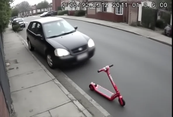 CCTV footage still of a car speeding away from a curb with a child's scooter lying on the pavement.